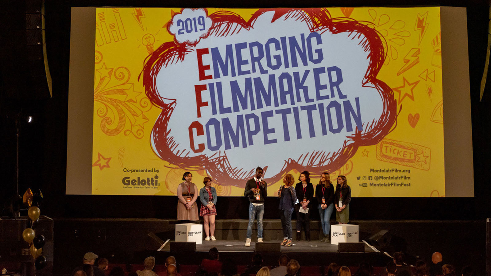 A group of students stand on stage in front of a large screen that says, 