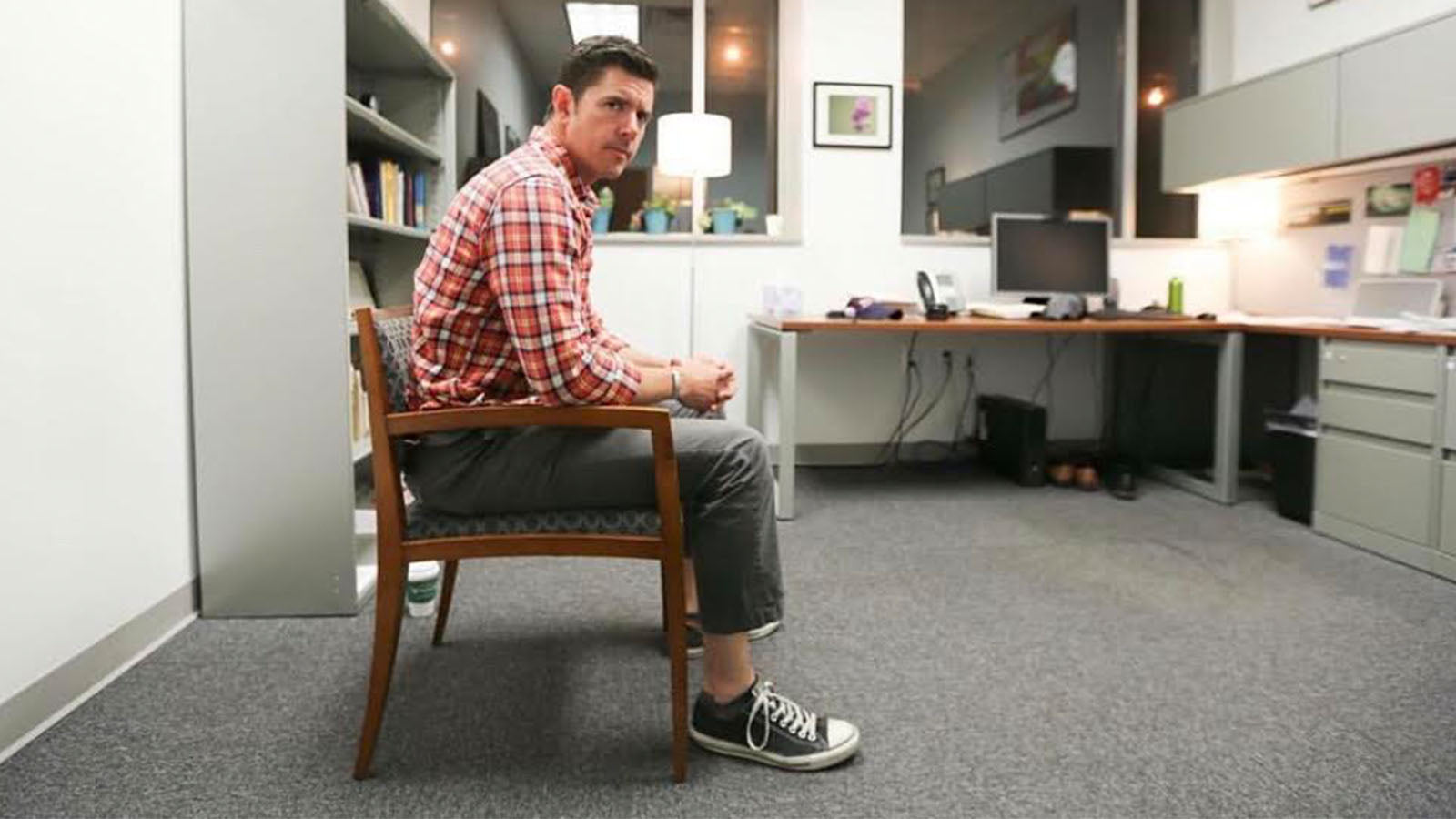 A man in a plaid shirt sits in a chair in an office.